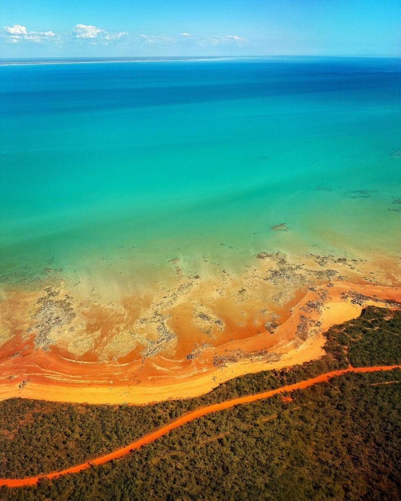 Aerial view of turquoise ocean meeting an orange sandy shore with a winding road through a dense green forest nearby.