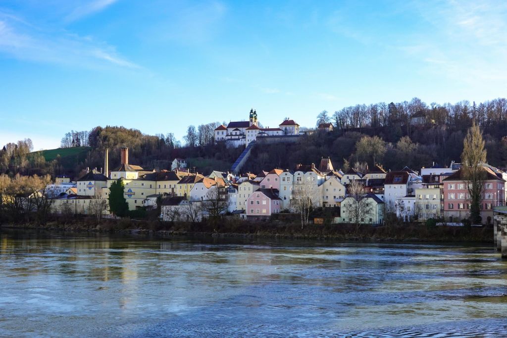 Colorful houses line a riverfront with a hill and church in the background under a clear blue sky