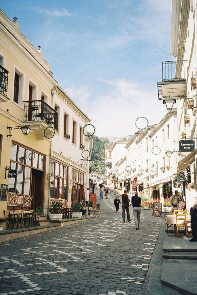 Pedestrian street in a European town with yellow and white buildings, outdoor cafes, and people walking on a cobblestone road under a blue sky.