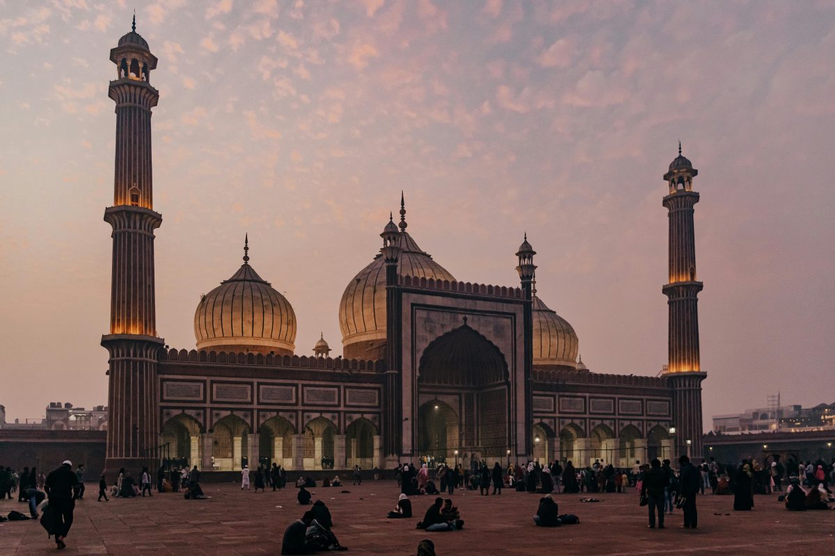 Sunset over a grand mosque with tall minarets and domed roofs, people gathered in the large courtyard.