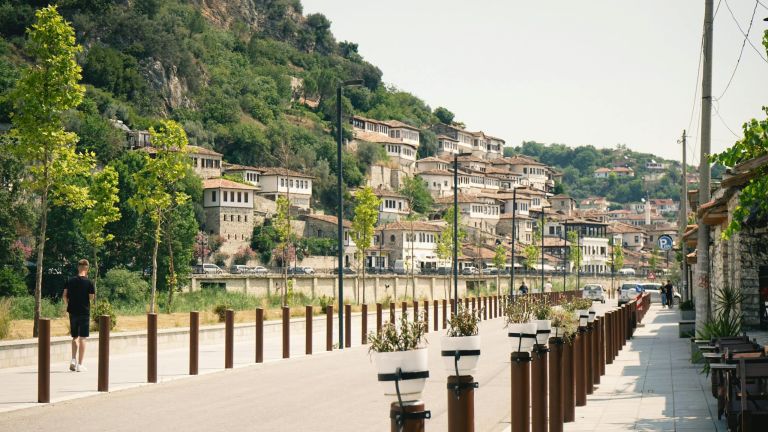 A man in a black shirt walks along a tree-lined promenade with brown bollards and a hillside town in the background.