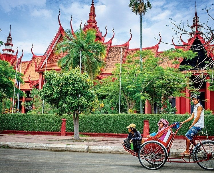 Cycle rickshaw with two passengers and a driver on a street in front of a red temple with ornate curved roofs and palm trees.