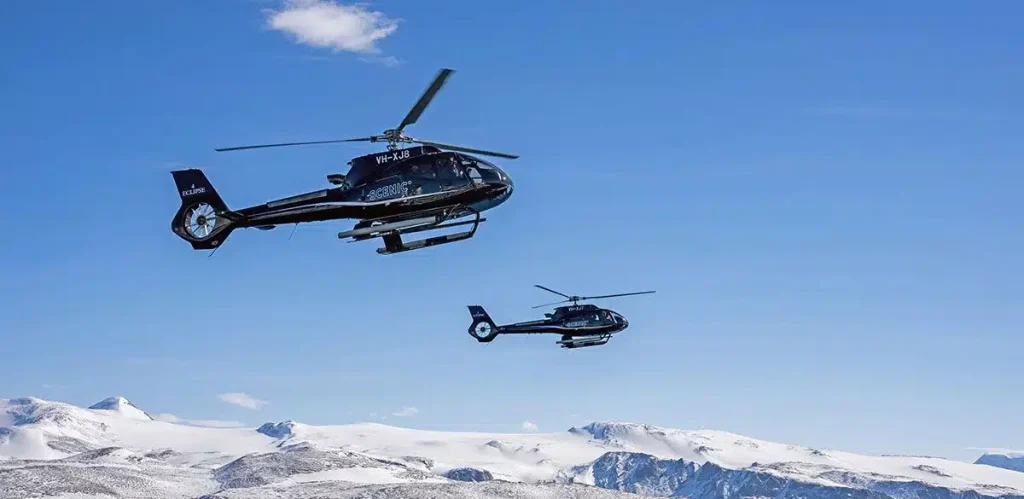 Two dark helicopters fly over a snow-covered mountain landscape under a clear blue sky, one ahead and closer to camera.