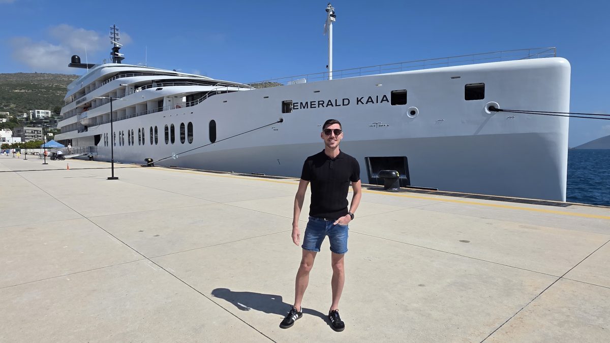 Man in black shirt and blue shorts poses on a dock beside a large white luxury yacht named Emerald Kaia.