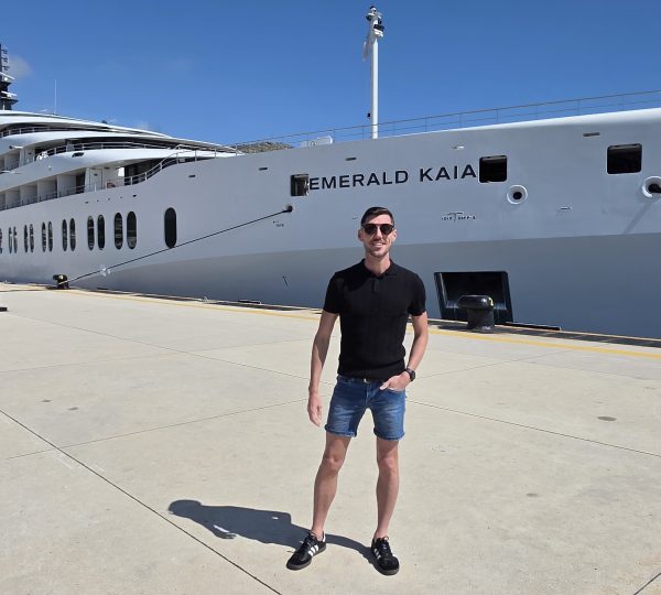 Man in black shirt and blue shorts poses on a dock beside a large white luxury yacht named Emerald Kaia.
