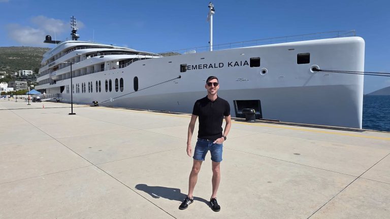 Man in black shirt and blue shorts poses on a dock beside a large white luxury yacht named Emerald Kaia.