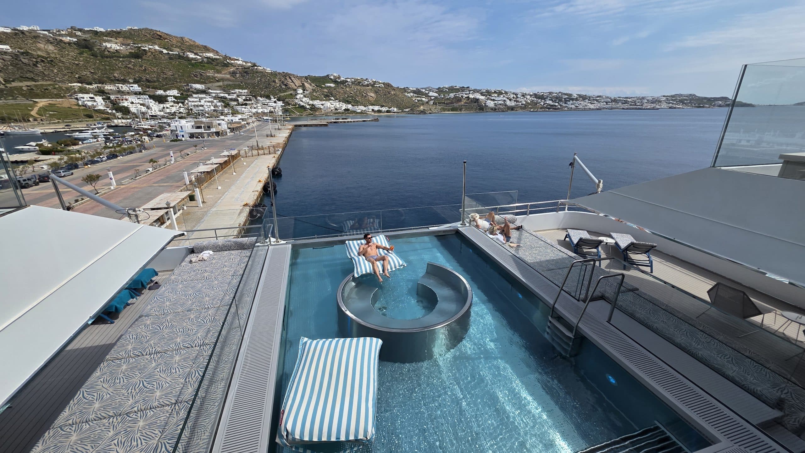 Relaxing hotel pool deck overlooking a coastal town and calm blue sea with people sunbathing and lounge chairs nearby