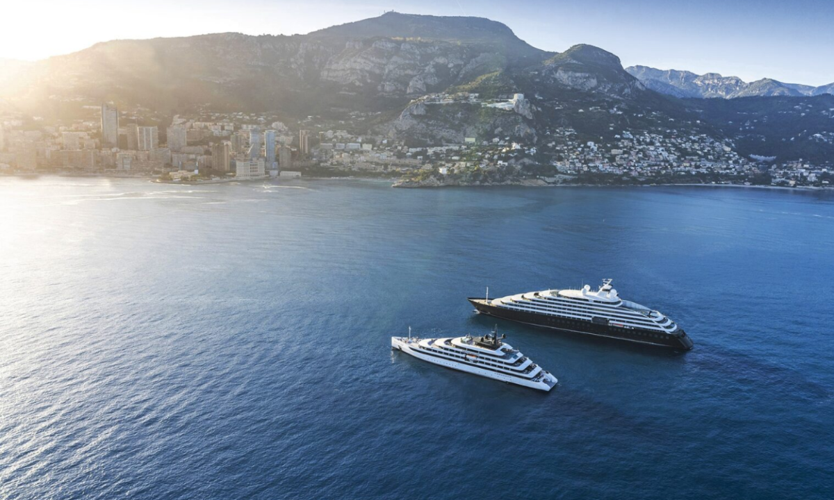 Aerial view of a coastal city with mountains in the background and two luxury yachts anchored in a calm blue harbor.