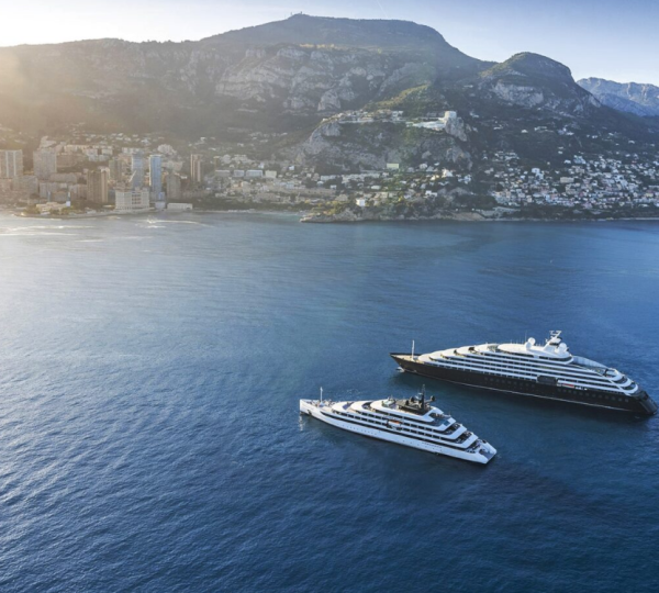 Aerial view of a coastal city with mountains in the background and two luxury yachts anchored in a calm blue harbor.