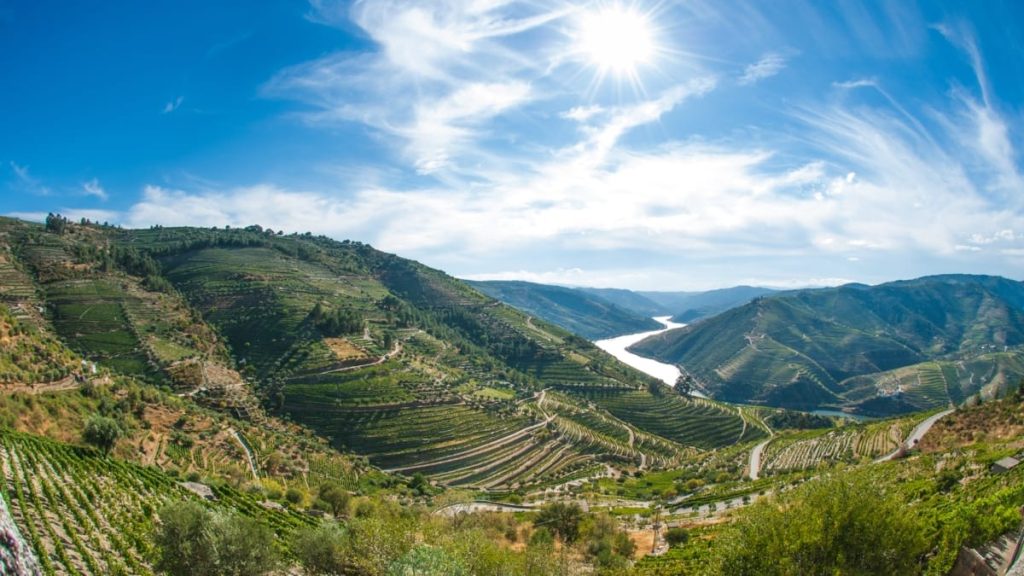 Terraced green hillsides with a winding river beneath a bright blue sky and sunburst.