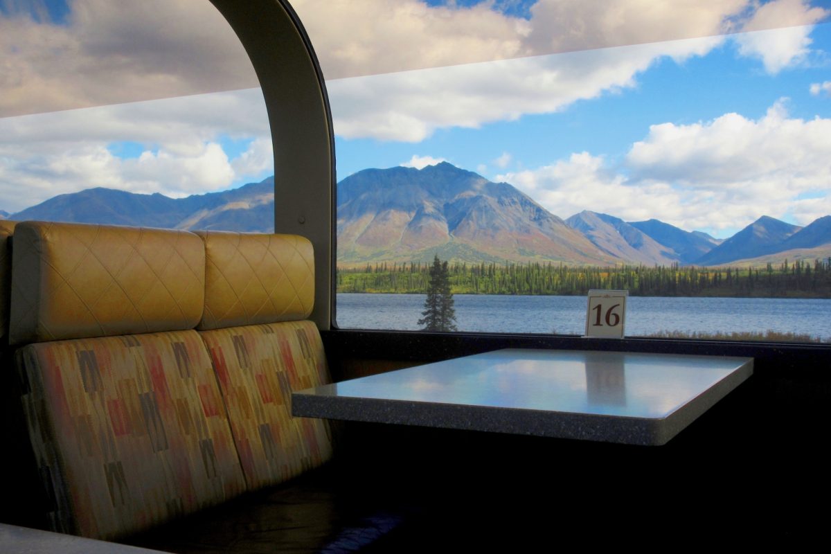 Bus interior with quilted tan seats and a table, view of a lake and mountains through a window; a small sign reading 16 on the sill. Alaska Railroad.