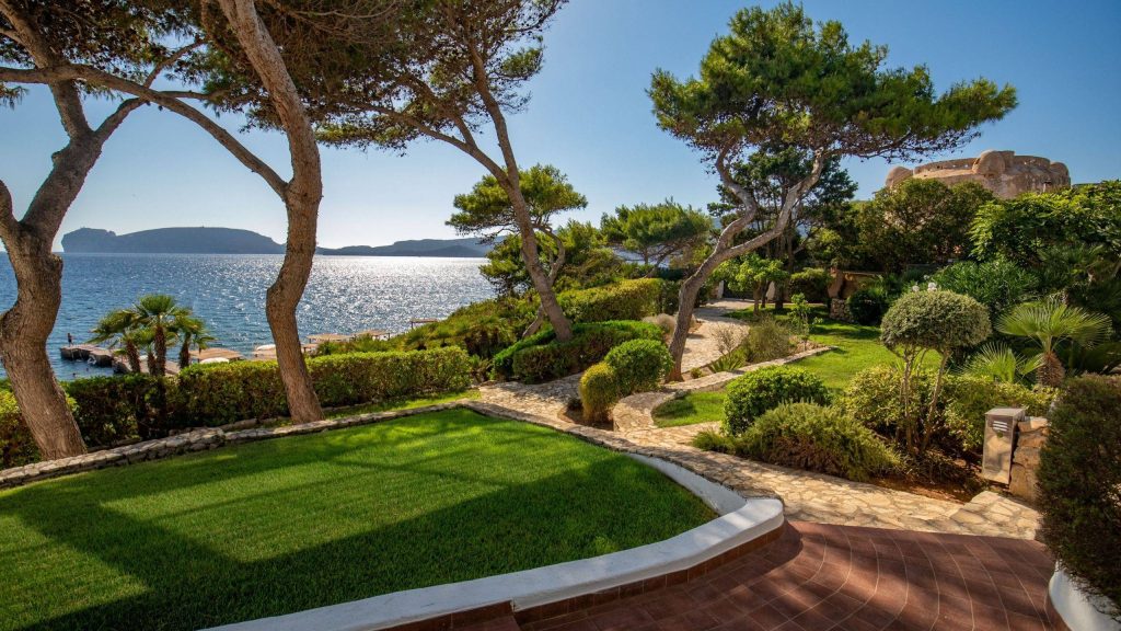 Sunlit seaside garden with a stone path, green lawn, and tall pine trees overlooking a blue bay and distant islands.