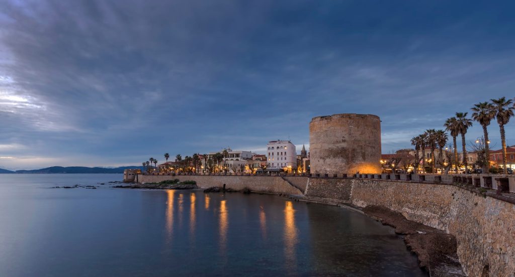 Circular stone tower along a seaside promenade at dusk, with palm trees and warm building lights reflecting on calm water.