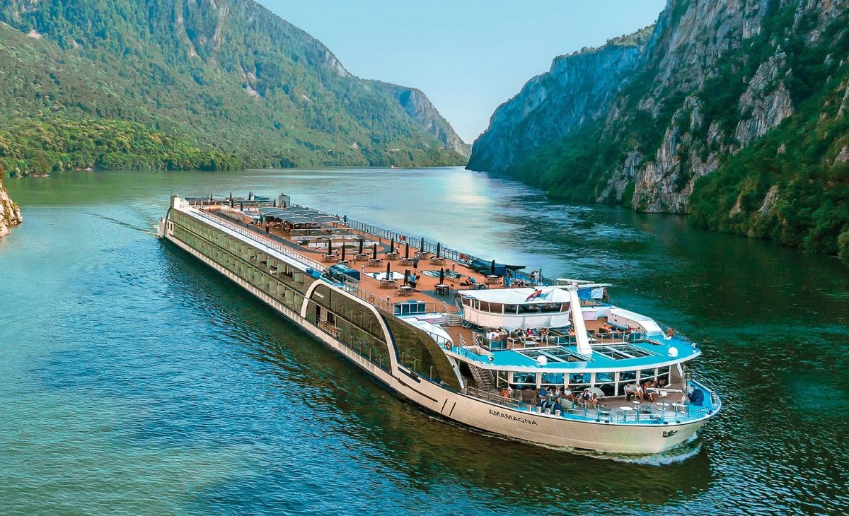 Large passenger cruise ship Ama Magna cruising on a turquoise river between steep, forested cliffs.