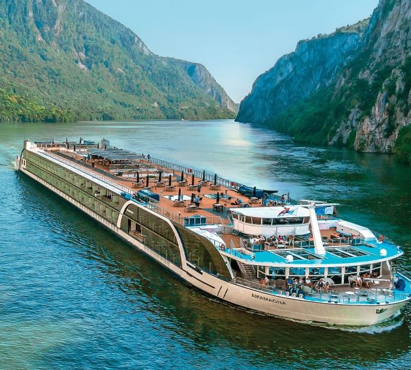 Large passenger cruise ship Ama Magna cruising on a turquoise river between steep, forested cliffs.