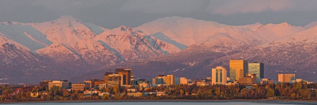 City skyline along a lake with snow‑capped mountains in the background bathed in warm sunset light, modern high‑rise buildings visible at shore.