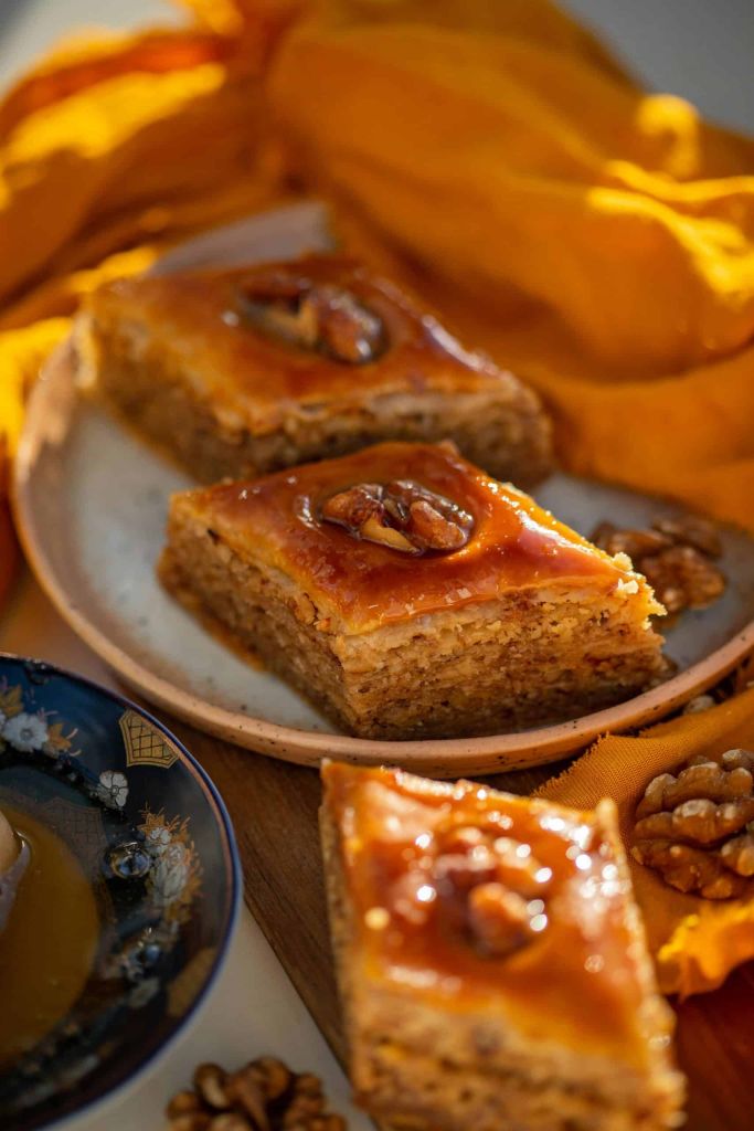 Slices of walnut-topped baklava with honey glaze on a ceramic plate beside orange fabric.