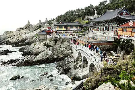 Temple complex perched on rocky coast with a white arched bridge and visitors walking along the path by the sea.