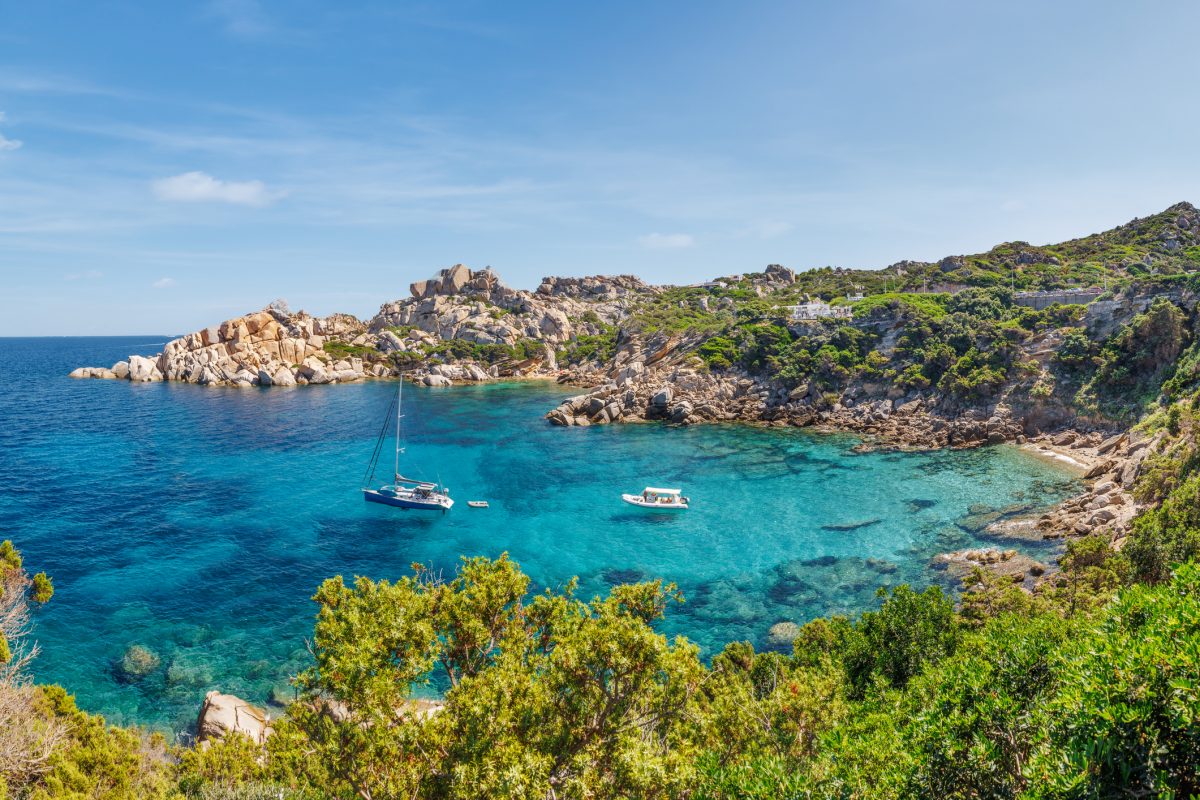 Turquoise bay with rocky coastline, green shrubbery, and small sailboats anchored in calm water under a blue sky in Sardinia