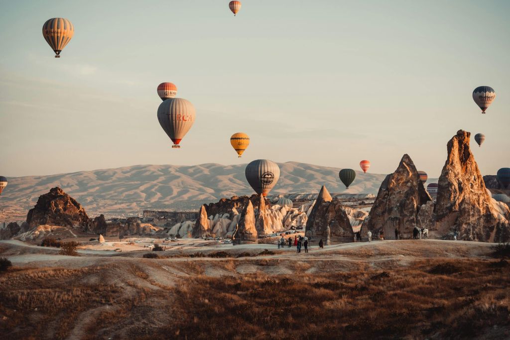 Multiple colorful hot air balloons rise over a rocky desert landscape with people strolling below at dusk/dawn.