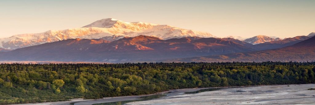 Snow-capped mountains glow at sunset over a green forest and winding river valley in the foreground.