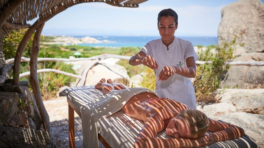 Therapist in a white uniform gives a back massage to a woman lying on an outdoor massage table by the sea under a wooden shade.