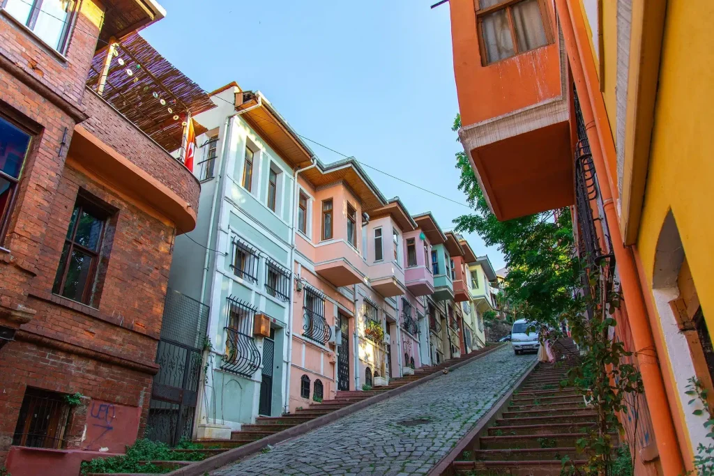 Colorful narrow street with pastel row houses and long stone stairs winding uphill under a clear blue sky, string lights overhead.