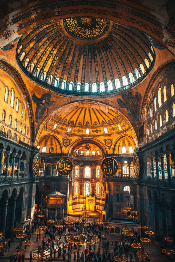 Interior of Hagia Sophia with a gigantic central dome, ornate arches, and hanging chandeliers, visitors fill the nave.