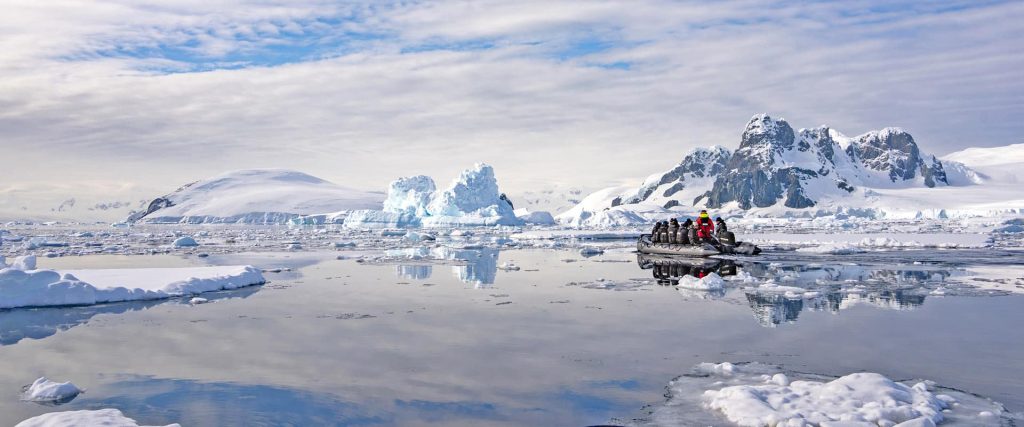 A team of explorers in a gray inflatable boat paddling through icy Arctic waters, with snow‑capped mountains and floating ice in the background.