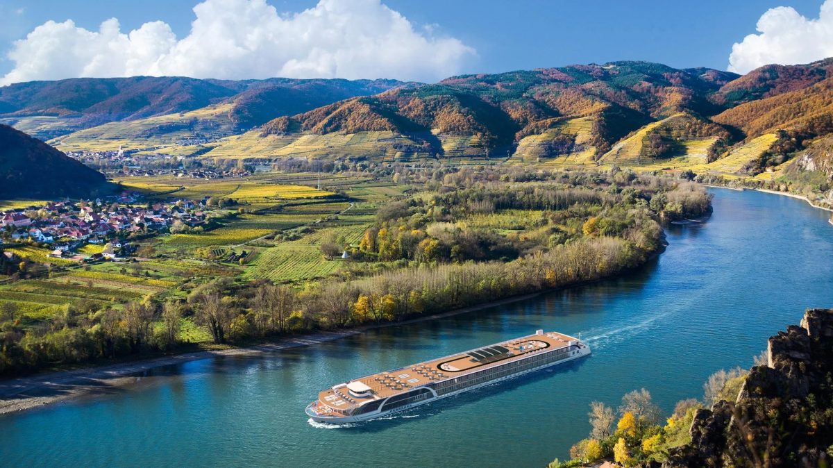 Large river cruise ship glides along a winding river through a terraced vineyard valley with forested hills under a blue sky.