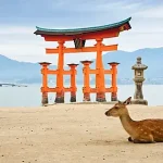 Deer resting on a sandy beach in front of a bright red torii gate by the water, with stone lanterns and distant mountains.