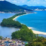 Aerial view of a curved sandy peninsula jutting into the blue sea, with a town along the shore and forested hills in the background.