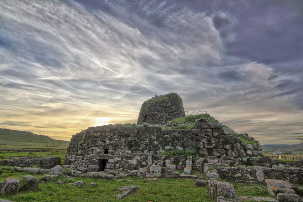 Circular stone ruin atop a low hill, with layered masonry and green vegetation, under a cloudy sunset sky