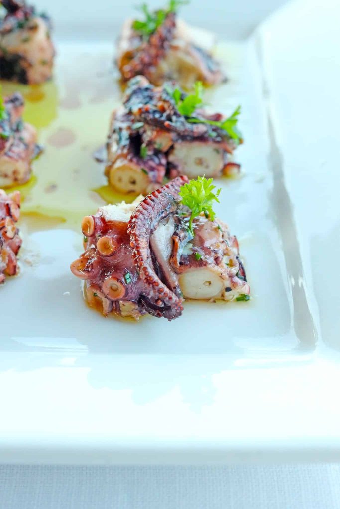 Sliced octopus appetizer on a white plate, garnished with parsley and herbs, drizzled with sauce.