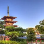 Three-story red pagoda with blue-tiled roofs in a traditional temple garden, surrounded by trimmed trees under a clear blue sky