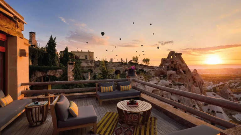 Sunset view from a stone terrace with cozy seating and a distant village; hot air balloons rise in the sky. Note: This alt text describes the scene and mood for someone viewing at sunset with balloons.