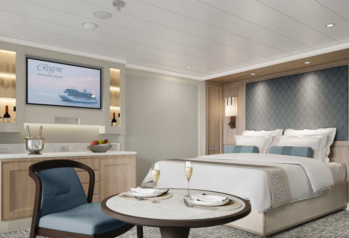 Luxurious cruise cabin with a king bed, seating area, and round table set for two with champagne glasses; TV screen shows Regent Seven Seas Cruises.
