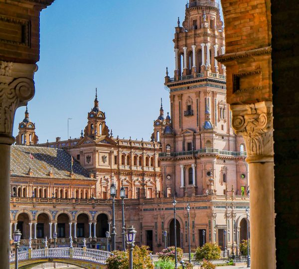 Sunlit Plaza de España with tall brick towers, ornate arches, and a decorative bridge over a canal.