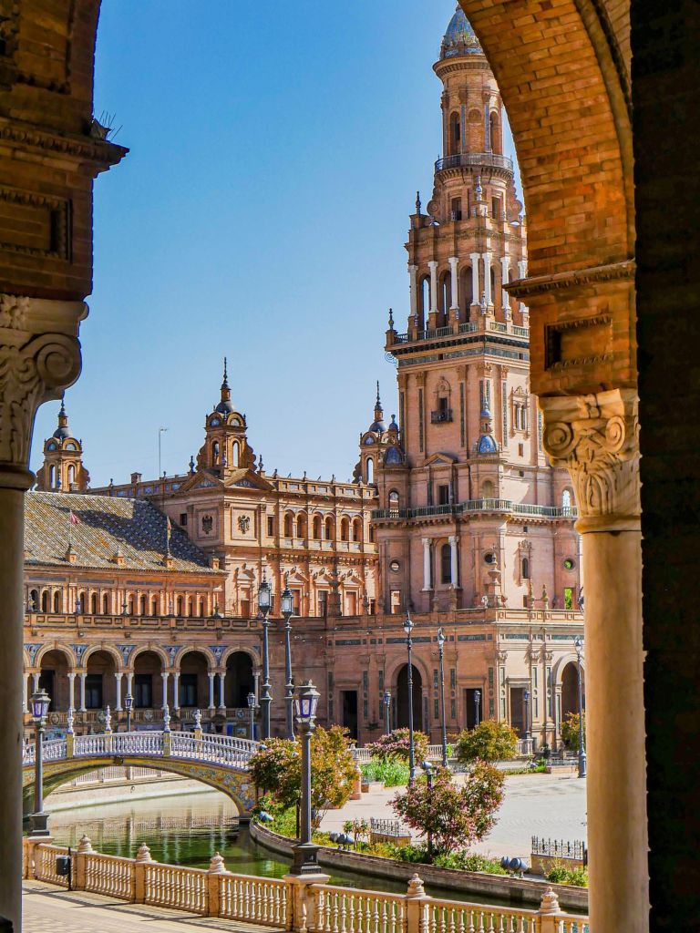 Sunlit Plaza de España with tall brick towers, ornate arches, and a decorative bridge over a canal.