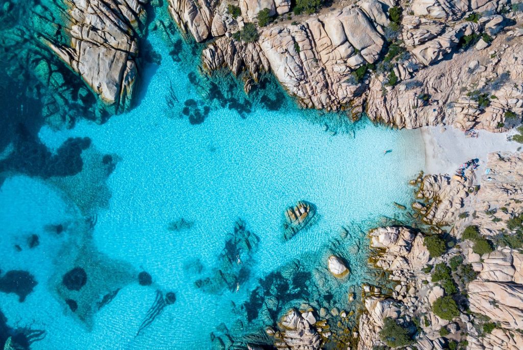 Aerial view of clear turquoise water meeting a rugged rocky shoreline with a small sandy beach and people at the beach area