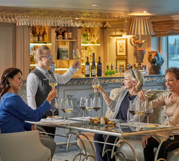 Four women toast with wine around a glass-topped table in a luxurious bar, smiling at a server standing nearby.