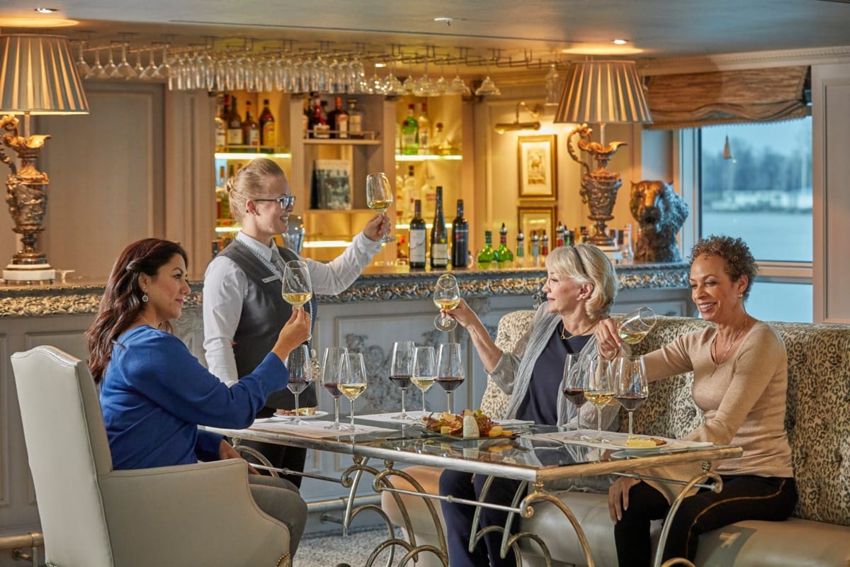 Four women toast with wine around a glass-topped table in a luxurious bar, smiling at a server standing nearby.