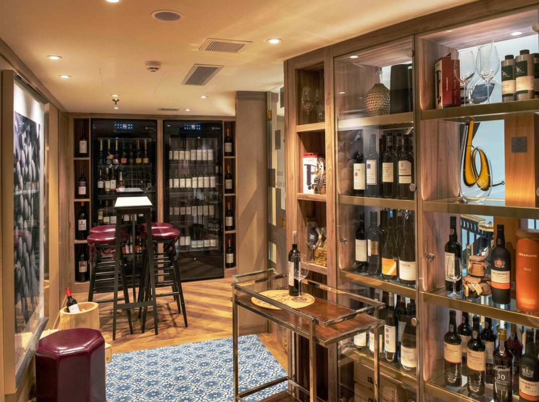 Cozy wine cellar with glass-front wine racks, bottled wines, and a small table with two burgundy stools in the foreground.