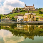 Castle on a hill overlooking a riverside town with pastel houses and vineyard terraces, reflected in the calm river.