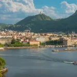 Panoramic view of a European lakeside city with historic buildings, a domed church, and green hills in the background; a boat moves across calm water in the foreground.
