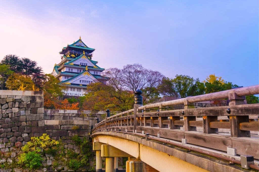 Japanese castle with green-tiled roofs sits on stone walls, viewed from a curved wooden bridge with autumn trees nearby.