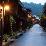 Narrow, wet street in a traditional Japanese village at dusk, lined with wooden houses and warm lantern light. Mountaintops are visible in the background.