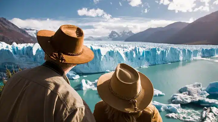 Two people wearing brown hats stand at the edge of a glacial lagoon, looking out at a blue glacier and snow-capped mountains.