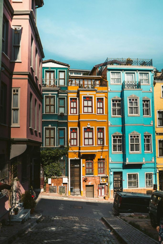 Colorful row of pastel houses (pink, teal, orange) along a narrow cobblestone street under a blue sky, with a black car parked on the right side and balconies visible.