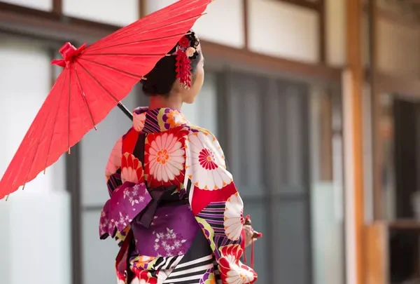 Woman in a colorful floral kimono seen from behind, holding a red parasol outside a traditional building area.